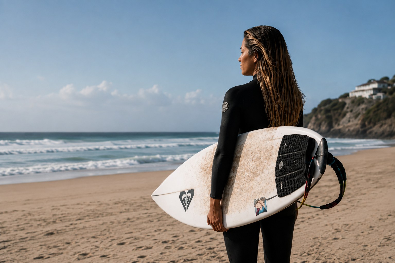 Surfista de costas na praia, segurando a prancha e olhando o mar em um momento de pausa e reflexão